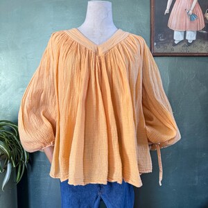 May include: A peach-colored blouse with a V-neck and long, gathered sleeves. The blouse has a textured fabric and is displayed on a mannequin. The sleeves have ties at the cuffs. The blouse is paired with blue jeans.