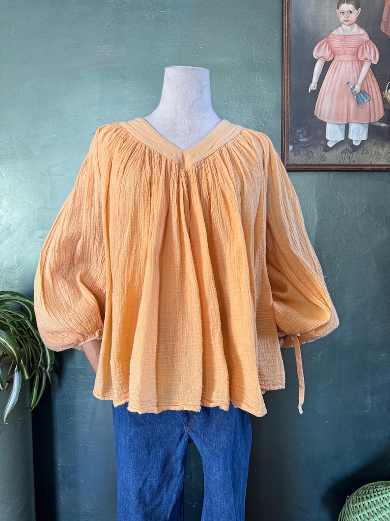 May include: A peach-colored blouse with a V-neck and long, gathered sleeves. The blouse has a textured fabric and is displayed on a mannequin. The sleeves have ties at the cuffs. The blouse is paired with blue jeans.