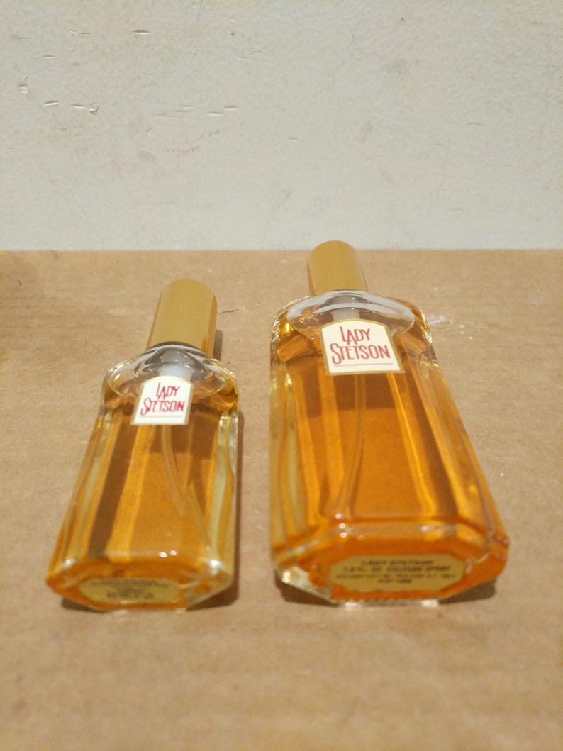 May include: Two glass bottles of perfume with gold caps. The bottles are clear and contain a yellow liquid. The bottles have a label that says "Lady Stetson" in red letters.