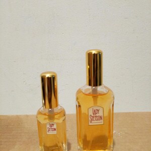 May include: Two glass bottles of Lady Stetson perfume. The smaller bottle is on the left and the larger bottle is on the right. Both bottles have gold caps and are filled with a light yellow liquid.