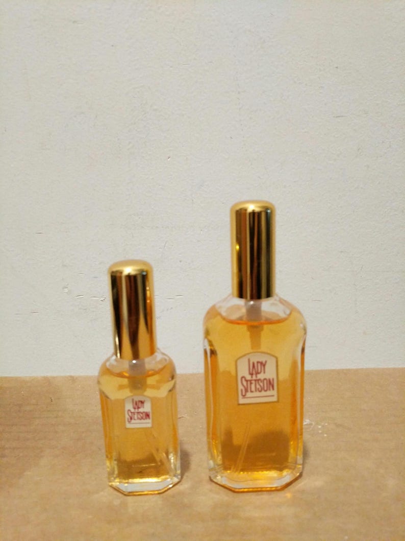 May include: Two glass bottles of Lady Stetson perfume. The smaller bottle is on the left and the larger bottle is on the right. Both bottles have gold caps and are filled with a light yellow liquid.