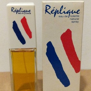 May include: A bottle of Réplique eau de toilette with its matching box. The bottle and box are white with blue and red brushstroke designs. The bottle contains a yellow liquid and is labeled "60 ML 2 FL OZ."