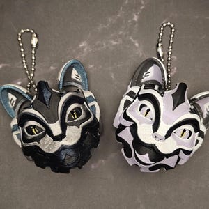 May include: Two cat-shaped keychains. One is black and gray with blue accents, the other is white, black, and lavender. Both have silver chain attachments and yellow eyes. The keychains are detailed and three-dimensional.