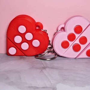 May include: Two heart-shaped keychains, one red and one pink, each with six circular accents. The red keychain has white accents, while the pink one has red accents. Both are attached to silver keyrings.