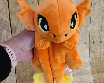 Puede incluir: Dragón de peluche naranja y amarillo con grandes ojos amarillos. El dragón está sentado con las alas extendidas.