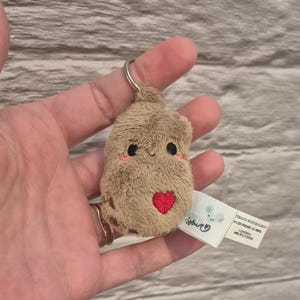 May include: A brown plush keychain shaped like a potato with a red heart embroidered on its chest. The potato has a smiling face with black eyes and a small mouth. The keychain is attached to a silver ring.