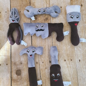 May include: A selection of soft toy tools, including a spanner, saw, hammer, screwdriver, pliers, and paintbrush. Each toy is grey and brown with embroidered faces. The toys are designed to look like they are smiling.