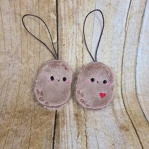 May include: Two brown plush potato keychains with embroidered faces and a red heart on one. The keychains have black strings.