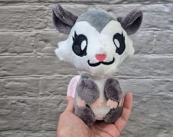 May include: A plush opossum toy with a white face, gray ears, and gray and pink paws. The toy has large black eyes, a pink nose, and a smiling mouth. The stuffed animal is held in a hand.