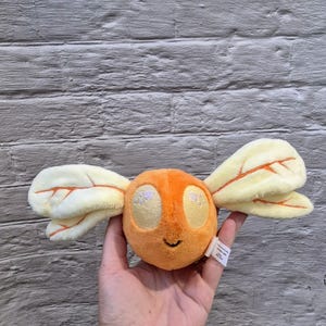 May include: A plush toy bee with an orange body, yellow eyes, and cream-coloured wings with orange vein details. The bee has a small, smiling mouth. The toy is held in a hand against a grey brick background.