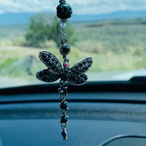 May include: A green and silver car charm featuring a dragonfly with crystal accents. The dragonfly is hanging from a beaded chain with a green and silver bead at the top.