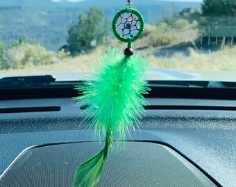 May include: Green dreamcatcher with a green feather hanging from the rearview mirror of a car. The dreamcatcher has a beaded string and a green bead.