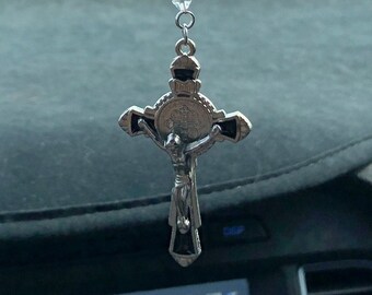 May include: A silver and black car charm with a crucifix and a beaded chain. The charm is hanging from the rearview mirror of a car.