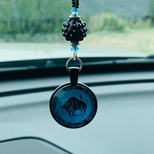 May include: A blue and black car charm with a black crystal bead and a round glass charm featuring a black silhouette of a bull. The charm is hanging from a rearview mirror.