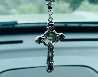 May include: A silver cross with a figure of Jesus Christ on it hangs from a black beaded chain with a silver bead at the top. The cross is hanging from a rearview mirror in a car.