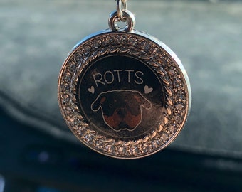 Op de afbeelding: Een zilverkleurige auto-hanger met een ronde hanger met het woord "ROTTS" en een Rottweiler-hondenillustratie. De hanger is omgeven door een ring van heldere kristallen. De hanger bevat decoratieve kralen en een zwart koord om op te hangen.