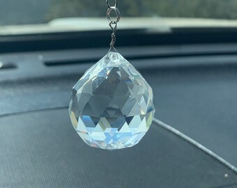 Puede incluir: Un amuleto de coche de cristal con un cristal transparente en forma de lágrima que cuelga de una cadena plateada con una pequeña perla de cristal iridiscente.