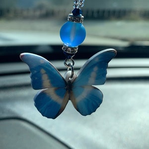 Puede incluir: Un amuleto de mariposa azul con un patrón azul claro y blanco cuelga de una cadena de cuentas con una cuenta de vidrio azul claro. El amuleto está sujeto a un espejo retrovisor.