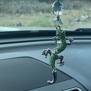 May include: A green jeweled lizard charm hanging from a rearview mirror. The lizard is made of metal and has a crystal bead hanging above it.