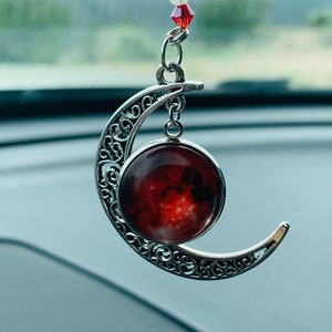 May include: A silver crescent moon charm with a red and white galaxy image inside. The charm is hanging from a black and white beaded chain with red and white beads.