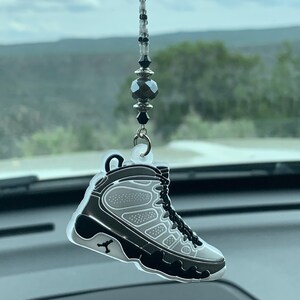 May include: A car charm featuring a black and gray sneaker design. The charm is suspended from a beaded strand with black and silver accents. The sneaker has a detailed design, including laces and the iconic Jumpman logo.