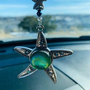 May include: A silver star-shaped car charm with a green iridescent shell-shaped center. The charm is hanging from a rearview mirror.