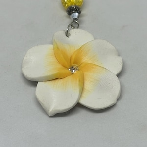 May include: A white and yellow plumeria flower pendant with a clear crystal center. The pendant is attached to a beaded strand with yellow and silver accents. The flower has a detailed, textured appearance.