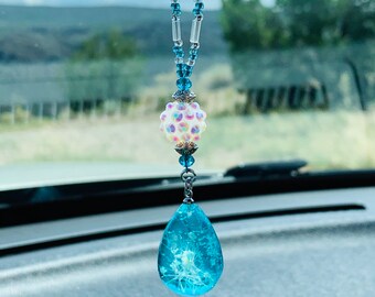 May include: A blue glass teardrop-shaped car charm with a white iridescent bead and silver accents hanging from a beaded chain.
