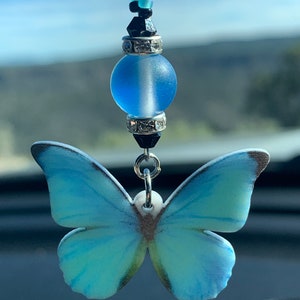 May include: A blue and white butterfly charm with a blue glass bead and black and blue beaded chain. The charm is hanging from a rearview mirror.
