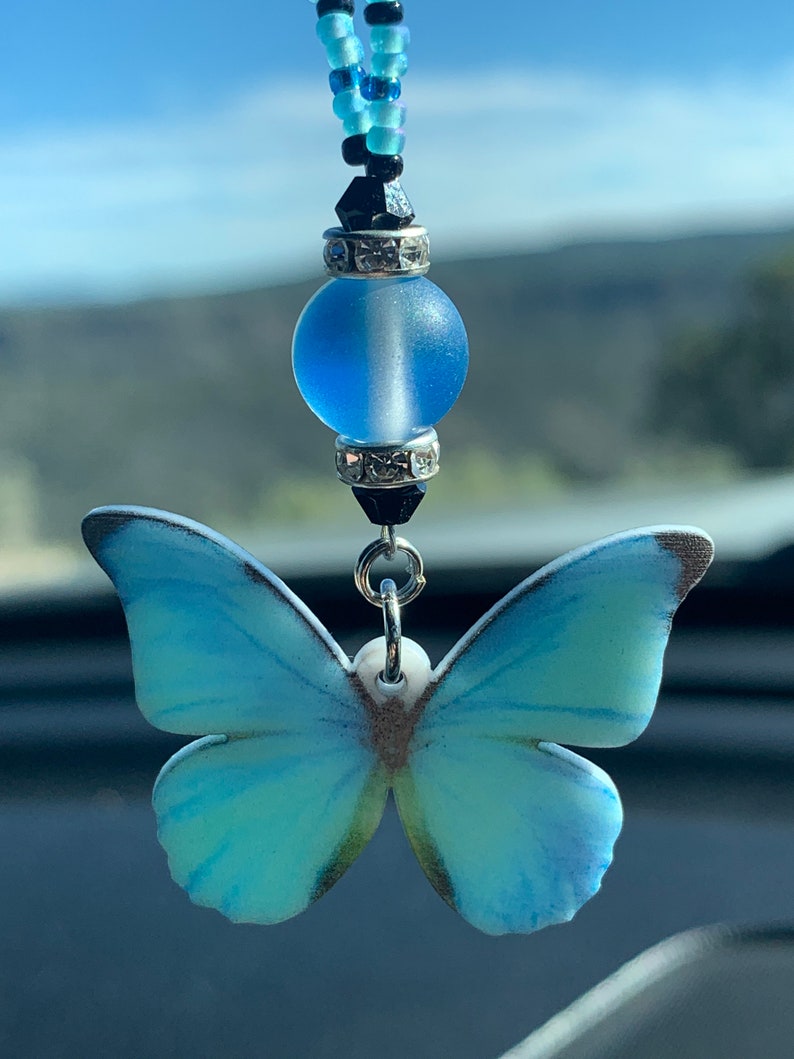 May include: A blue and white butterfly charm with a blue glass bead and black and blue beaded chain. The charm is hanging from a rearview mirror.