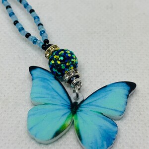 May include: A blue butterfly charm necklace with a sparkly green and black bead. The necklace is made of blue and black beads.