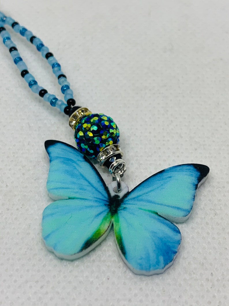 May include: A blue butterfly charm necklace with a sparkly green and black bead. The necklace is made of blue and black beads.