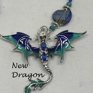 May include: A silver dragon charm with blue and green enamel wings and blue gem accents. The charm is attached to a beaded necklace.