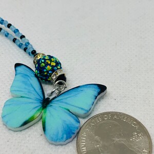 May include: A blue butterfly charm necklace with a sparkly green and black bead. The necklace is made of blue and black beads and is about the size of a US quarter.