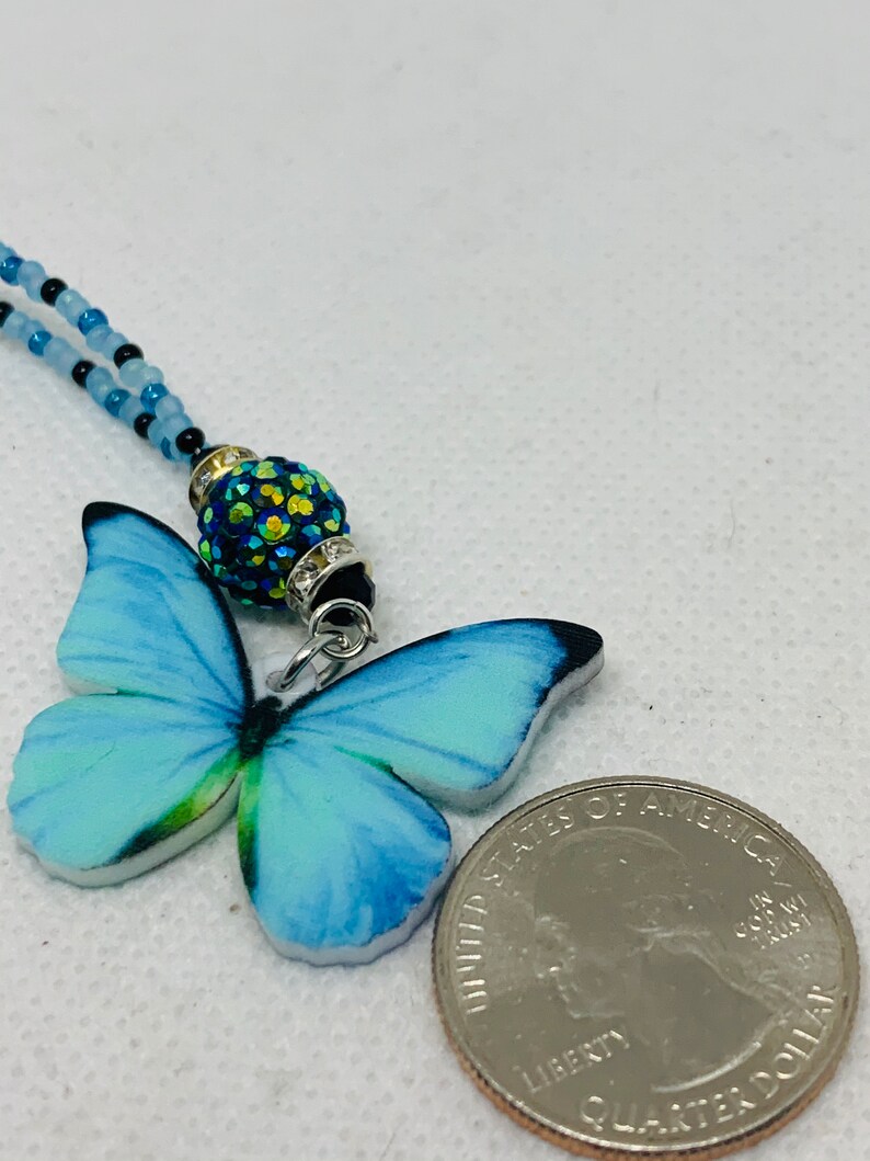 May include: A blue butterfly charm necklace with a sparkly green and black bead. The necklace is made of blue and black beads and is about the size of a US quarter.