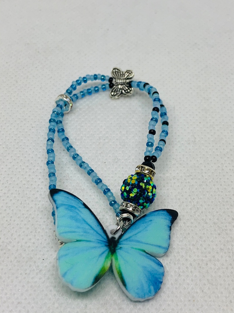 May include: A blue and black beaded bracelet with a butterfly charm and a sparkly green and black bead. The butterfly charm is silver and has a detailed design.