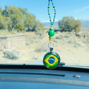 Puede incluir: Un amuleto para el espejo retrovisor con la bandera de Brasil. El amuleto está hecho de vidrio y metal y está enhebrado en una cadena de cuentas con cuentas verdes, azules y amarillas.