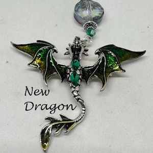 May include: A silver dragon pendant with green and yellow enamel wings and emerald-coloured accents. The dragon is suspended from a beaded strand with a large, iridescent bead. The text "New Dragon" is below the dragon.