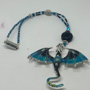 May include: A silver dragon pendant with blue and black enamel wings and a beaded cord. The dragon has a black body and is adorned with rhinestones. The cord features blue and black beads and a magnetic clasp.