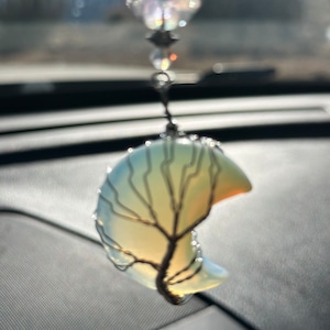 May include: A crescent moon-shaped crystal with a wire-wrapped tree design hangs from a rearview mirror. The crystal is translucent and has a light blue and yellow color.