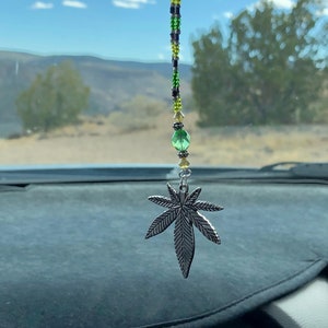 May include: A silver marijuana leaf charm hangs from a rearview mirror. The charm is attached to a beaded string with green, black and silver beads.