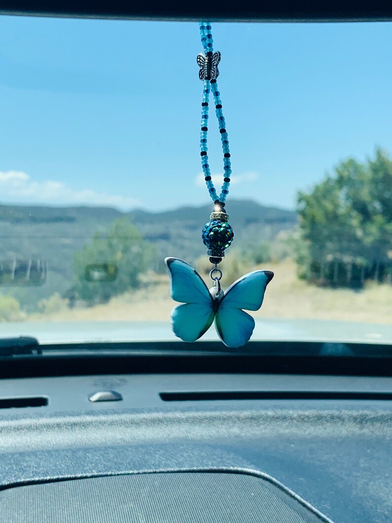 May include: A blue butterfly car charm with a beaded chain and a crystal bead. The charm is hanging from the rearview mirror of a car.