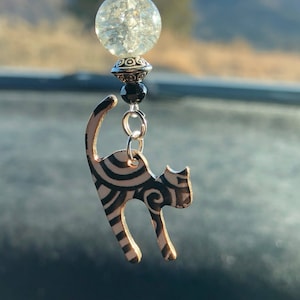 May include: A decorative car charm featuring a black and white cat pendant with a swirling design. The charm includes a clear, textured bead and black beads, suspended from a silver-toned chain. The background is blurred, suggesting an outdoor setting.
