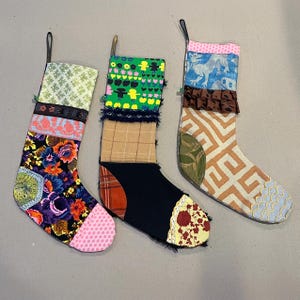 May include: Three colorful Christmas stockings made of patchwork fabric. The stockings are decorated with various patterns, including floral, geometric, and plaid designs. The stockings are hanging on a white wall.