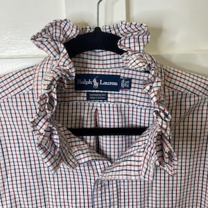 May include: A white long-sleeve button-down shirt with a red and navy blue grid pattern, featuring a ruffled collar. The interior label reads Ralph Lauren, Yarmouth, 100% Cotton, 17 1/2, 32/33. A small embroidered logo is visible on the lower front.