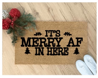 May include: A rectangular tan coir doormat featuring the black text IT'S MERRY AF IN HERE, accented with small black pine tree icons and decorative leaf flourishes. The mat is positioned near red and silver ornaments and green pine branches.