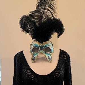 May include: A black dress with long sleeves and a scoop neckline, paired with a decorative mask and a feathered headpiece. The mask is teal, gold, and black. The dress has a fitted waist and a flared skirt.