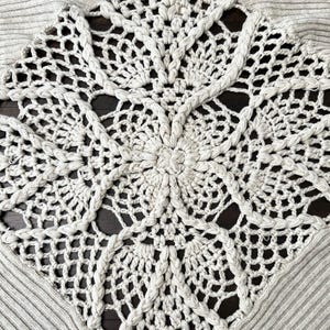 May include: A light grey sweater featuring a large, cream-coloured, crocheted square design on the front. The crocheted design has a central floral motif and intricate openwork patterns. The sweater has a ribbed texture.