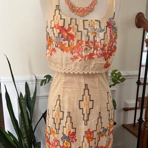 May include: A two-piece dress on a mannequin. The top is a square-neck tank with a floral print in orange, red, and purple. The skirt has a geometric pattern in beige and brown. A peach-colored beaded necklace is around the mannequin's neck.