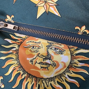 May include: Close-up of a dark teal fabric with a repeating pattern of gold stars, suns, and fleur-de-lis. A brass zipper bisects the image. The sun graphic is orange and yellow with a detailed face.
